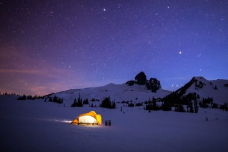 Glowing yellow tent with stars and Black Tusk in the background winter camping in Garibaldi Provincial Park near Whistler, BC.